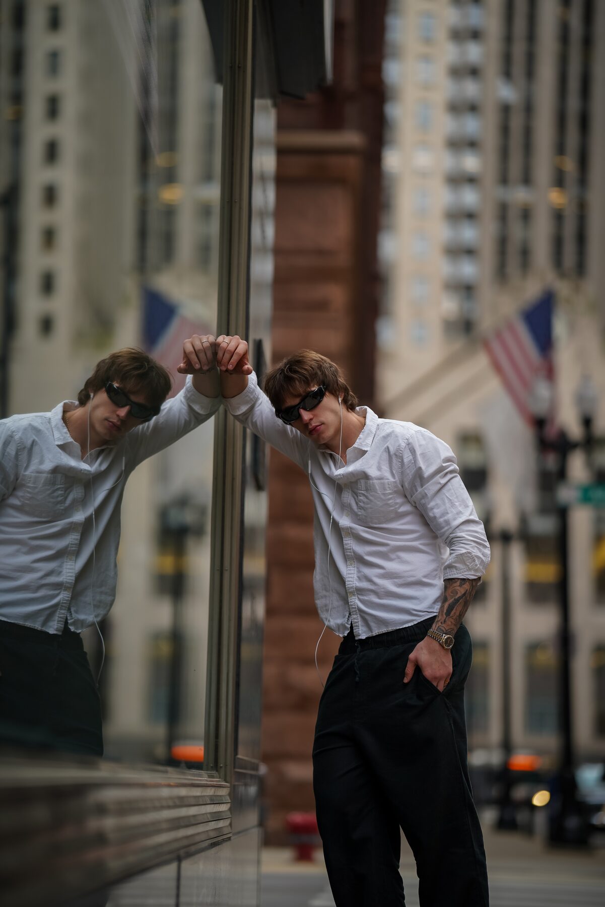 Sotos leans into a reflective storefront window, sunglasses on, an American flag and downtown architecture blurred in the background.