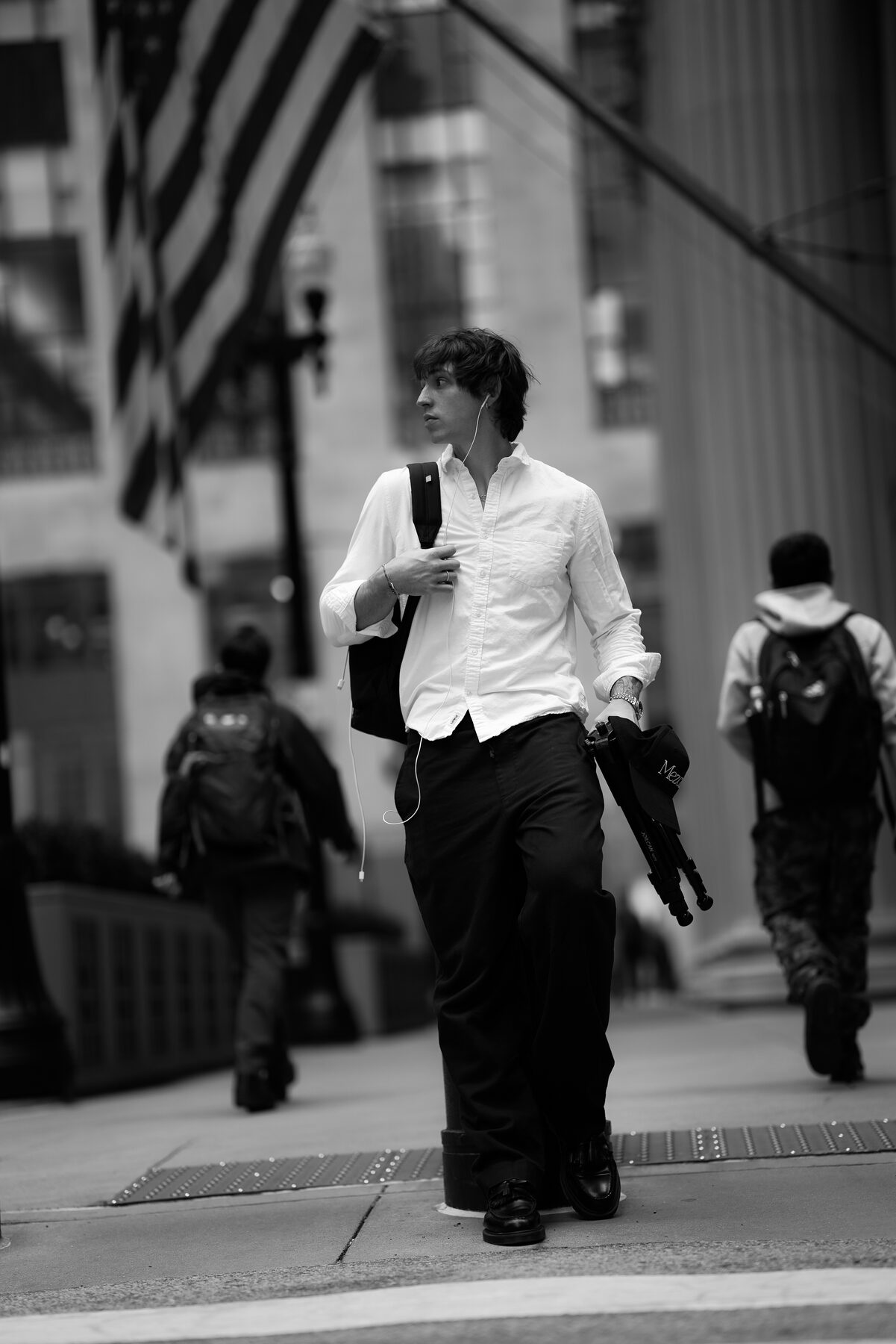 Sotos walking down a Chicago sidewalk with a backpack on one shoulder and a folded tripod in his hand — the means of production, carried by the maker.